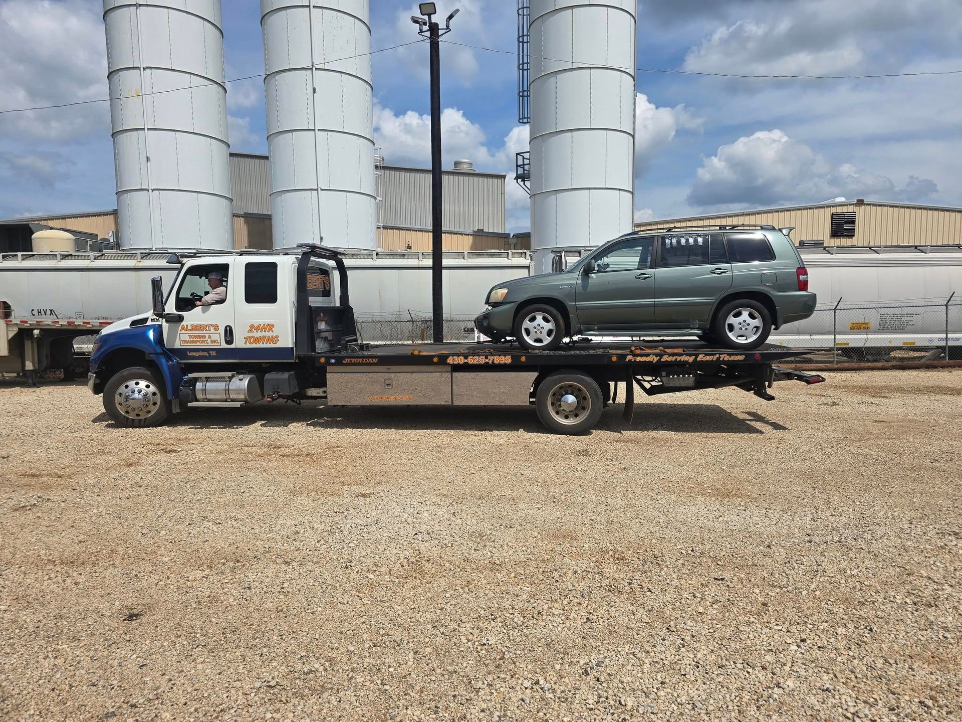 A tow truck with a green SUV on its flatbed, parked outdoors near silos.