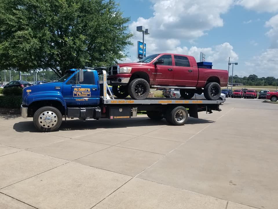 Blue tow truck carrying a red pickup truck on a sunny day at a dealership.