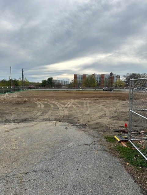 A fence surrounds a large dirt field with buildings in the background.