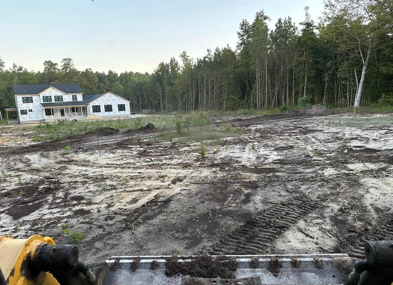 A person is driving a tractor through a muddy field with a house in the background.