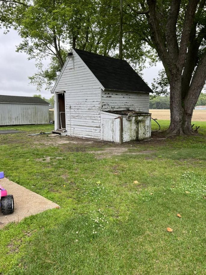 A small white shed is sitting in the middle of a grassy field next to a tree.