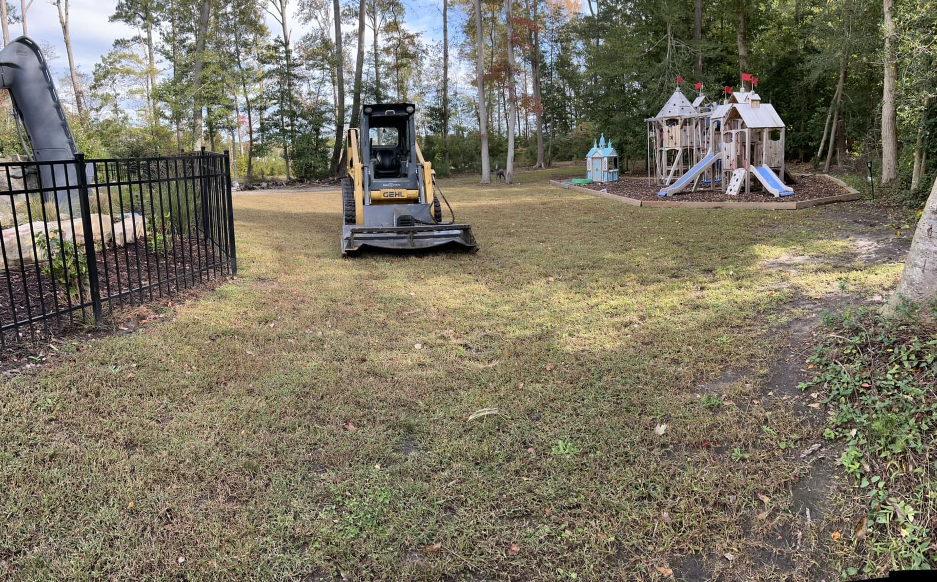 A bulldozer is cutting grass in a yard next to a playground.