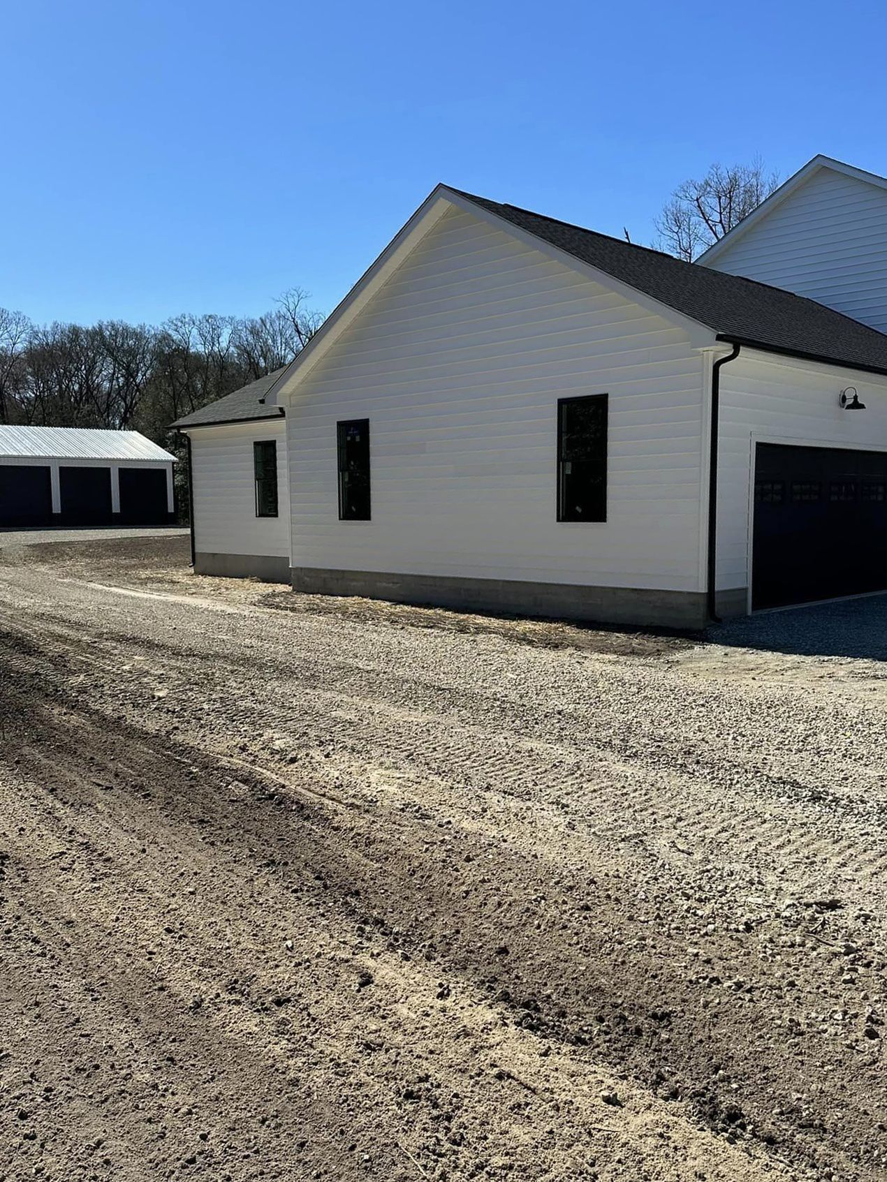 A white house with a black garage door is sitting next to a gravel road.