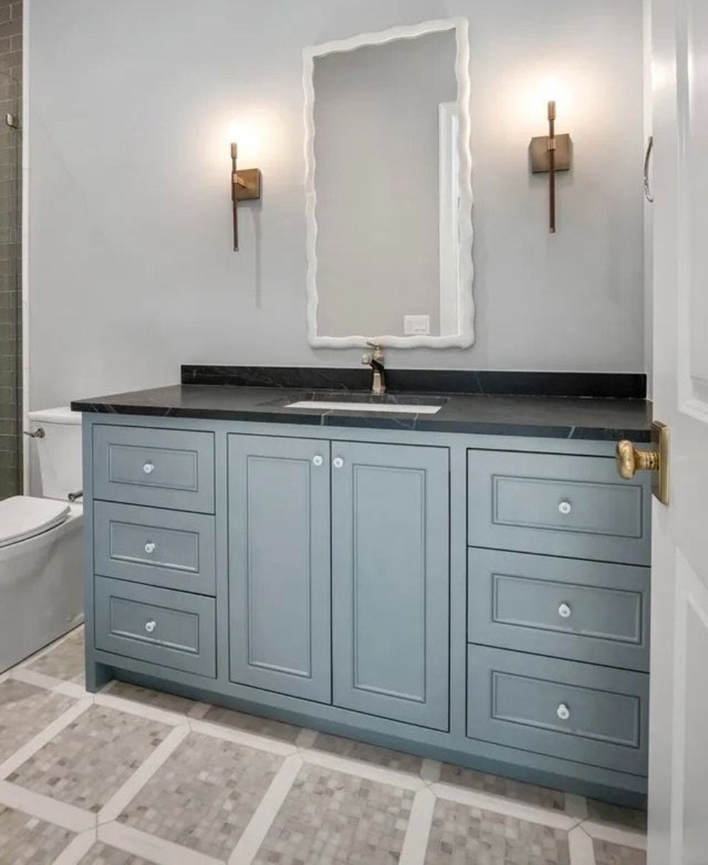 Light blue bathroom vanity with black countertop, gold fixtures, and mirror between wall sconces.