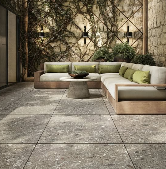 Modern stone patio with a beige sectional sofa, round table, and green cushions against a vine-covered wall.