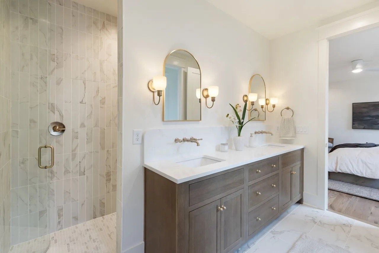 Bright bathroom with glass shower, double vanity, marble countertop, and mirror sconces