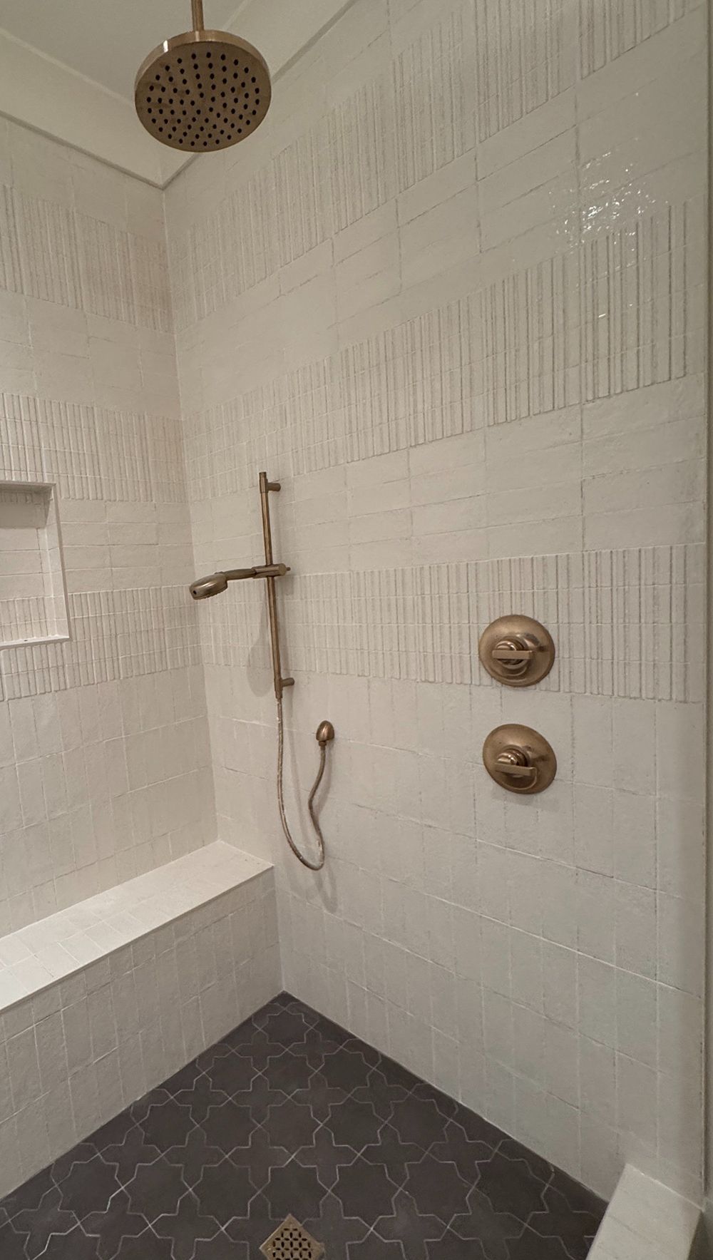 White tiled shower with dark hex floor, showerhead, handheld wand, two control knobs, and a bench seat.