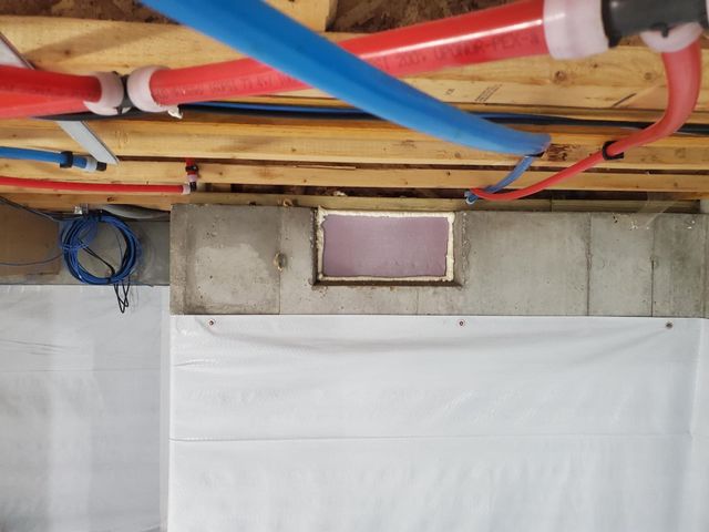 Basement wall with utility pipes overhead and a small pink vent opening near the ceiling