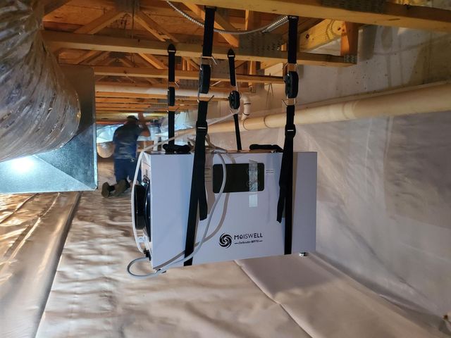 White HVAC unit suspended in an attic by black straps, with ductwork and insulation visible.