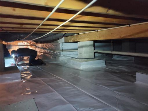 Worker crouching under a wooden structure, inspecting insulation in a dim crawlspace.