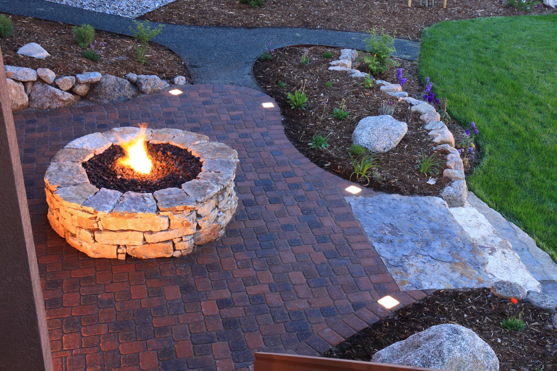 Stone fire pit burning on a brick patio beside landscaped rocks and grass
