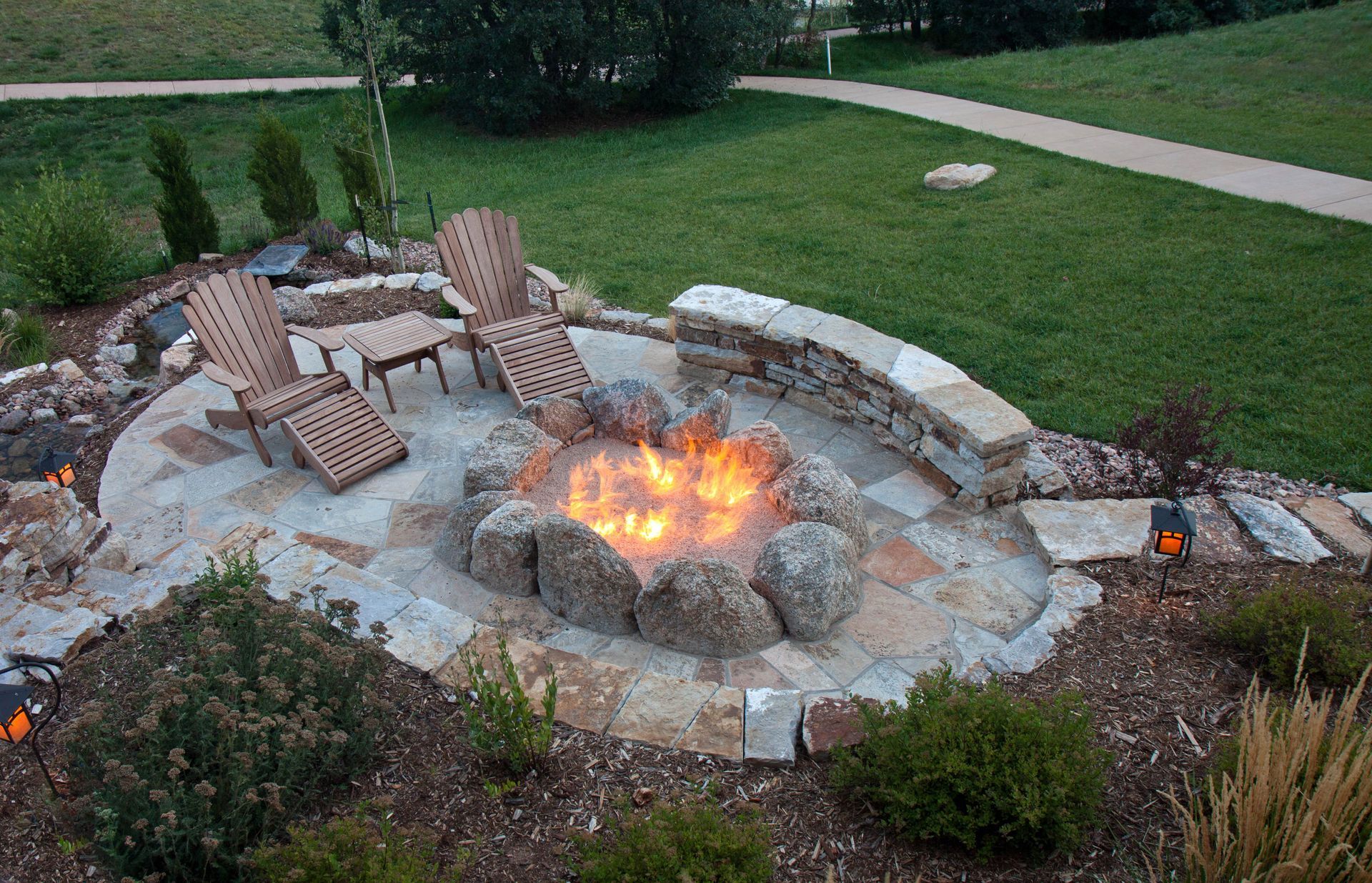 Stone backyard fire pit with flames, surrounded by Adirondack chairs and landscaping at dusk