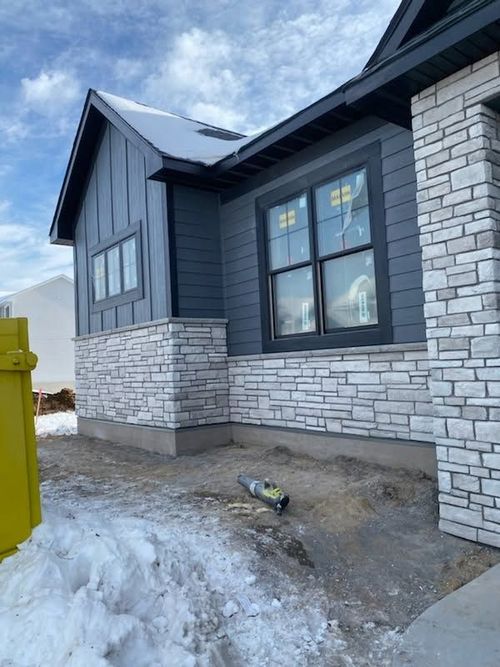 Snowy house exterior with stone and dark siding, windows, and a yellow bin by the front walkway