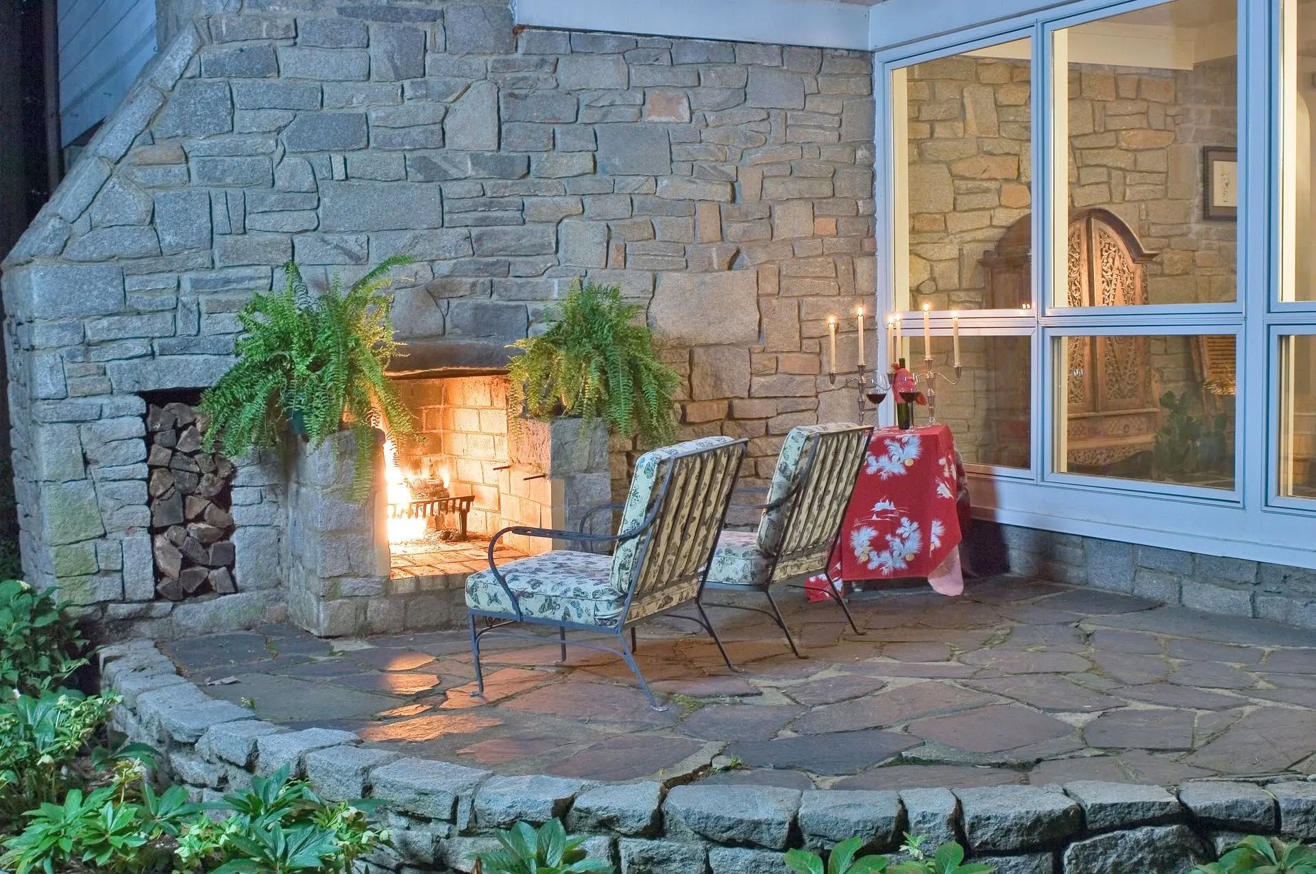 Stone patio with a lit outdoor fireplace, two chairs, potted plants, and a red side table by large windows