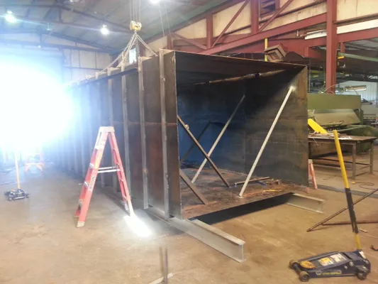 Large metal duct under construction in a workshop, with ladders, tools, and bright light glare on the left.