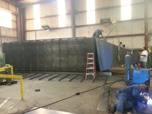 Workers welding a large metal structure inside a warehouse, with a red ladder and equipment nearby