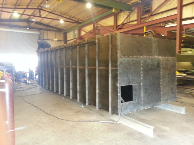 Large metal industrial enclosure inside a warehouse, with a small square opening and welded seams.