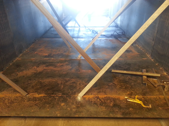 Inside a large metal container, wooden braces support a rust-stained floor under bright light.