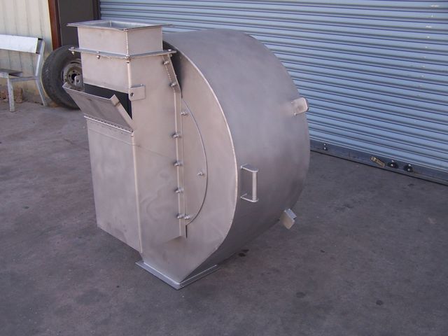 Stainless steel industrial machine with a curved housing, sitting on a concrete floor in front of a corrugated metal wall.