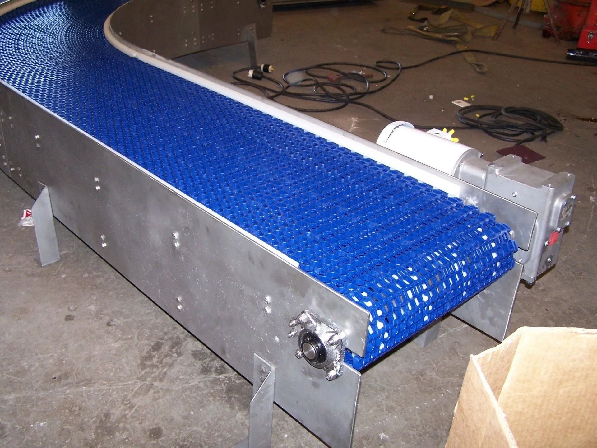 Blue conveyor belt on a metal frame in a workshop, with wiring and equipment nearby.