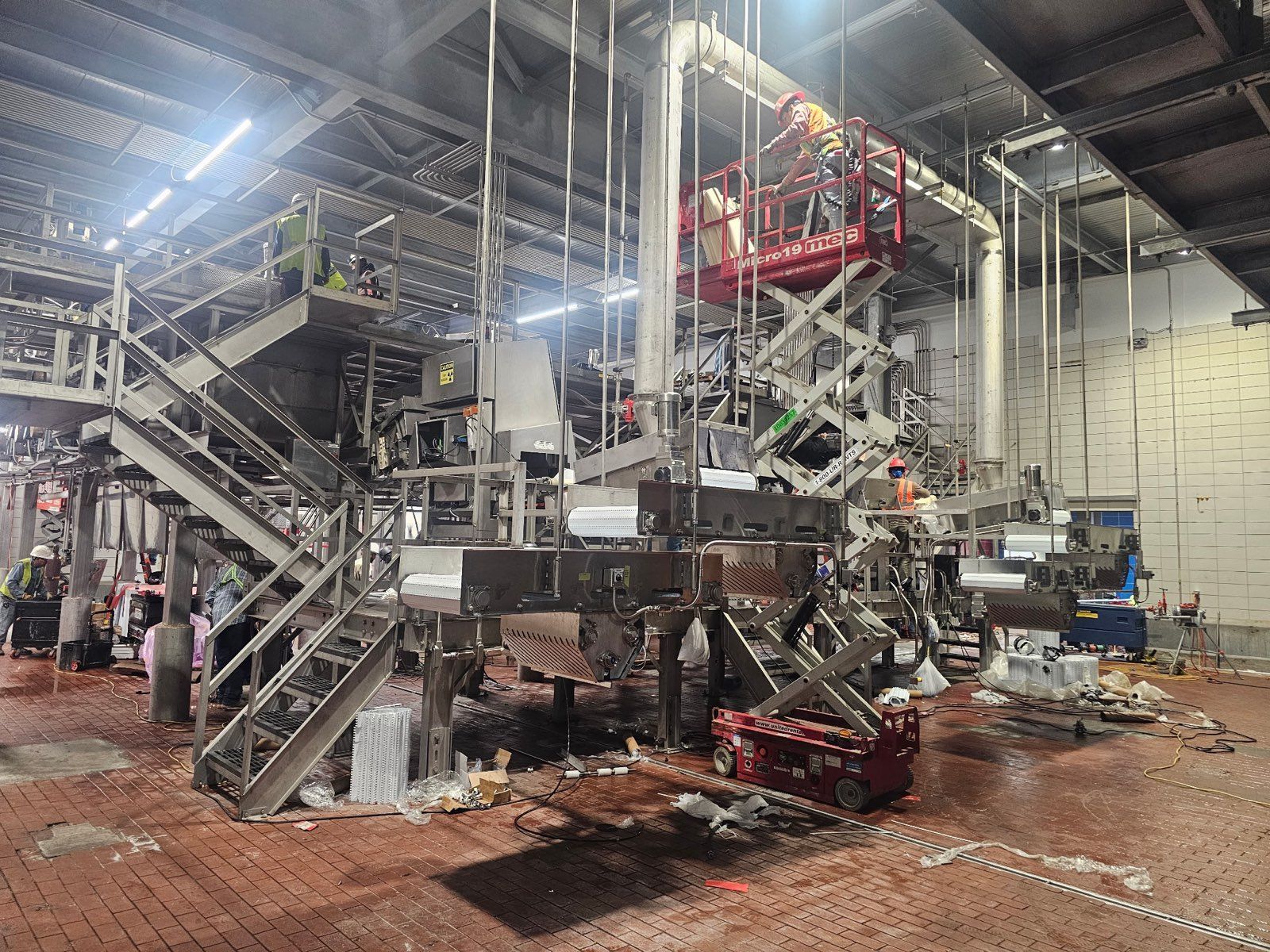 Large industrial machine with metal stairs and platforms in a warehouse-like factory, lit by bright overhead lights.