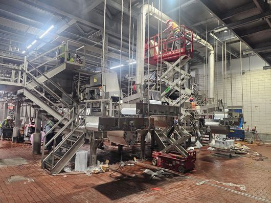Large industrial machine with metal stairs and platforms in a warehouse-like factory, lit by bright overhead lights.