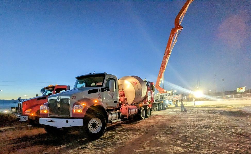 A concrete truck with a pump attached to it is driving down a dirt road at night.
