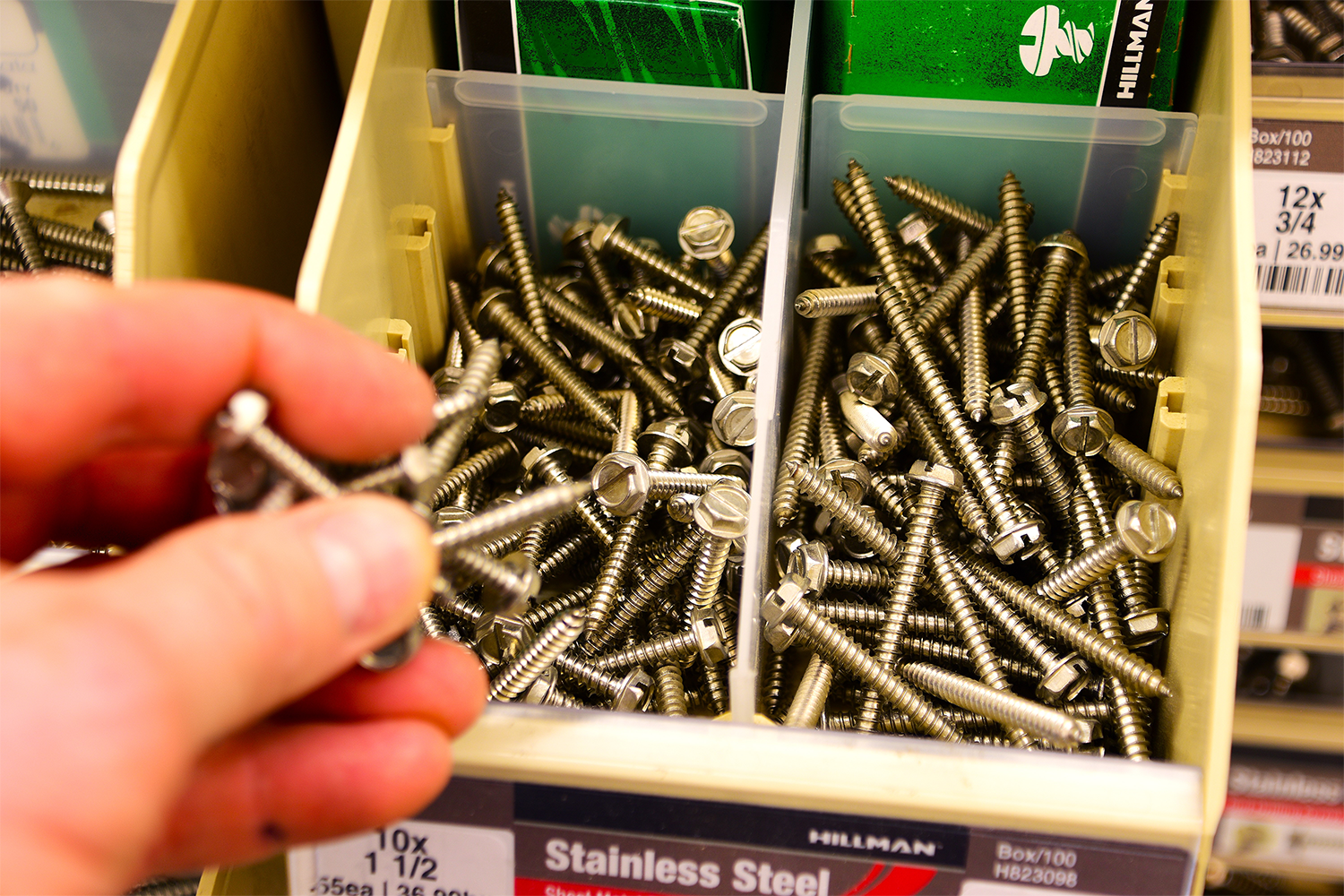 A person is holding a piece of stainless steel in front of a bin of screws.