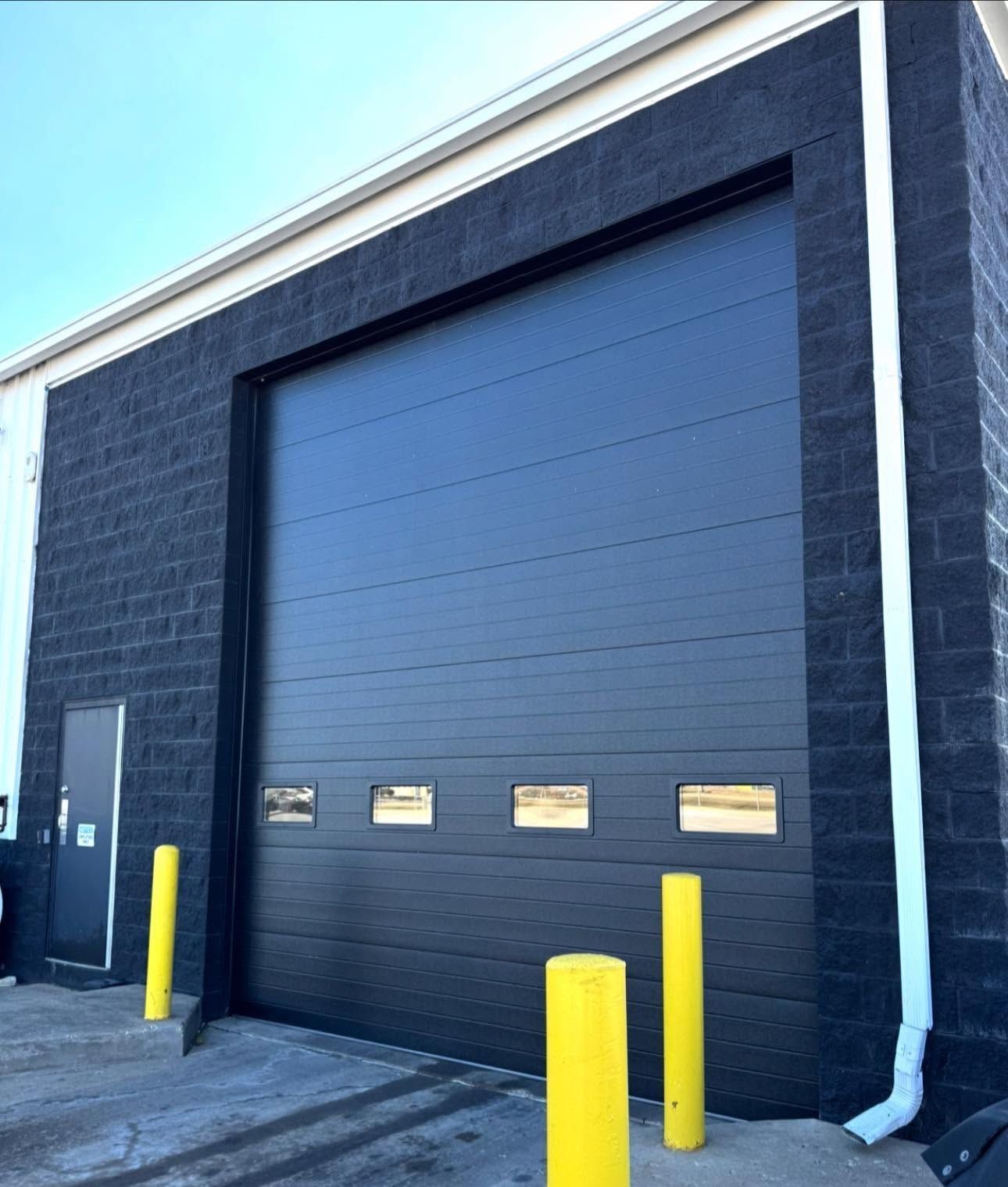 Black industrial garage door with small windows and yellow bollards outside a building