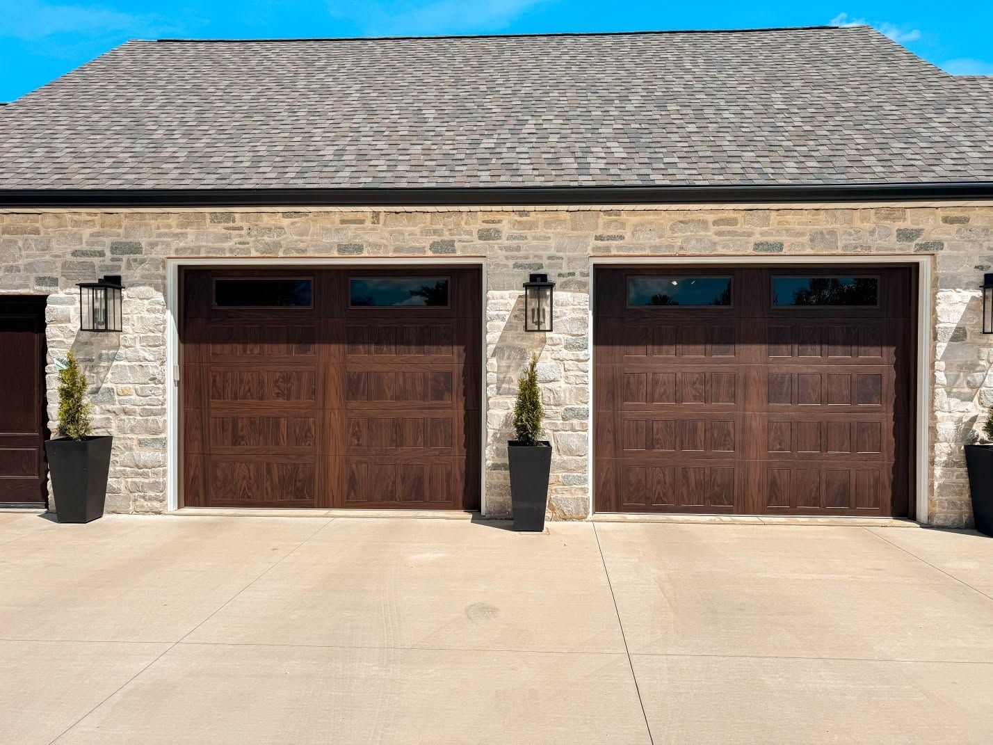Two dark wooden garage doors on a stone house with a light concrete driveway and potted plants