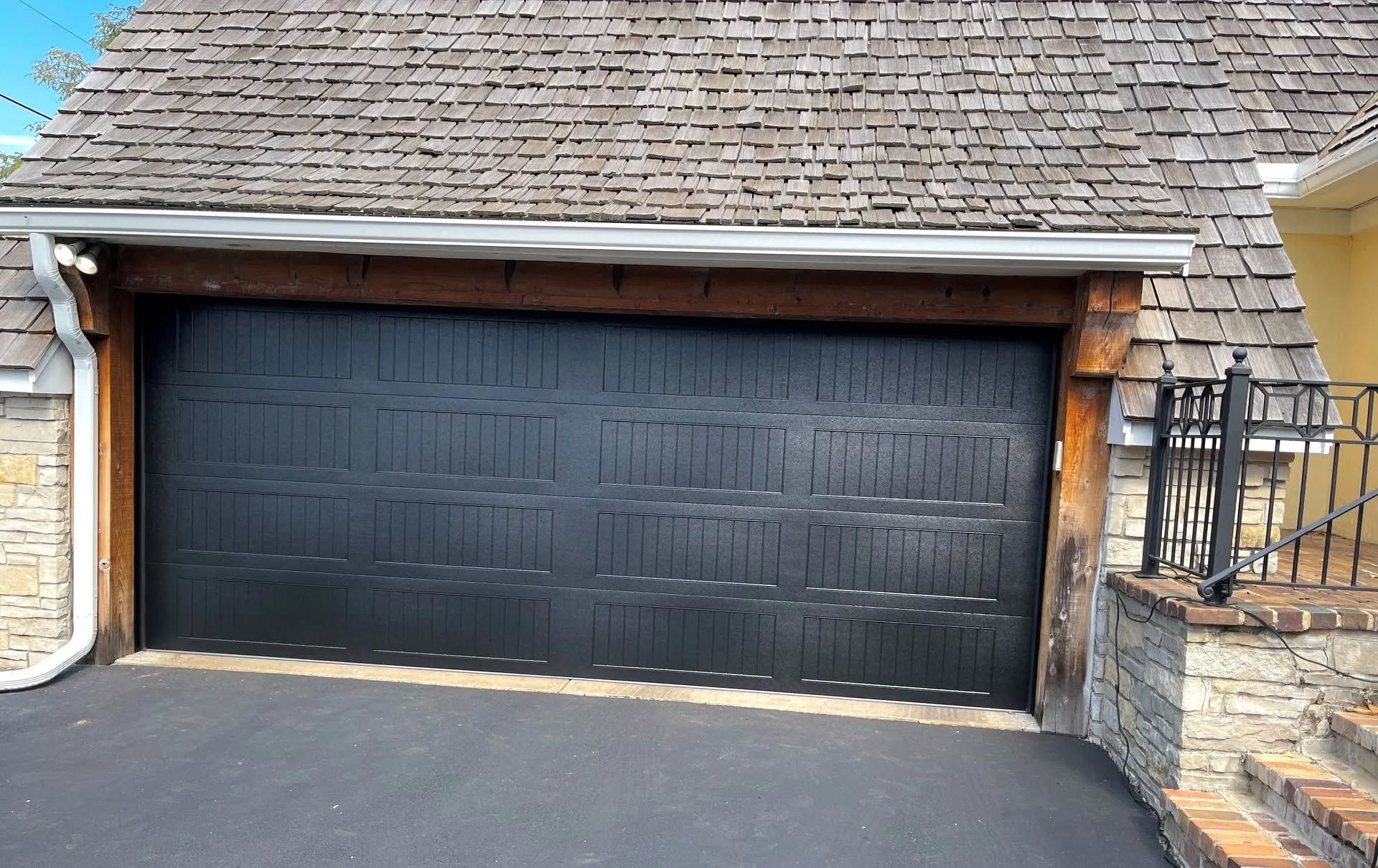 Black garage door under a shingled roof beside a stone and brick driveway entrance