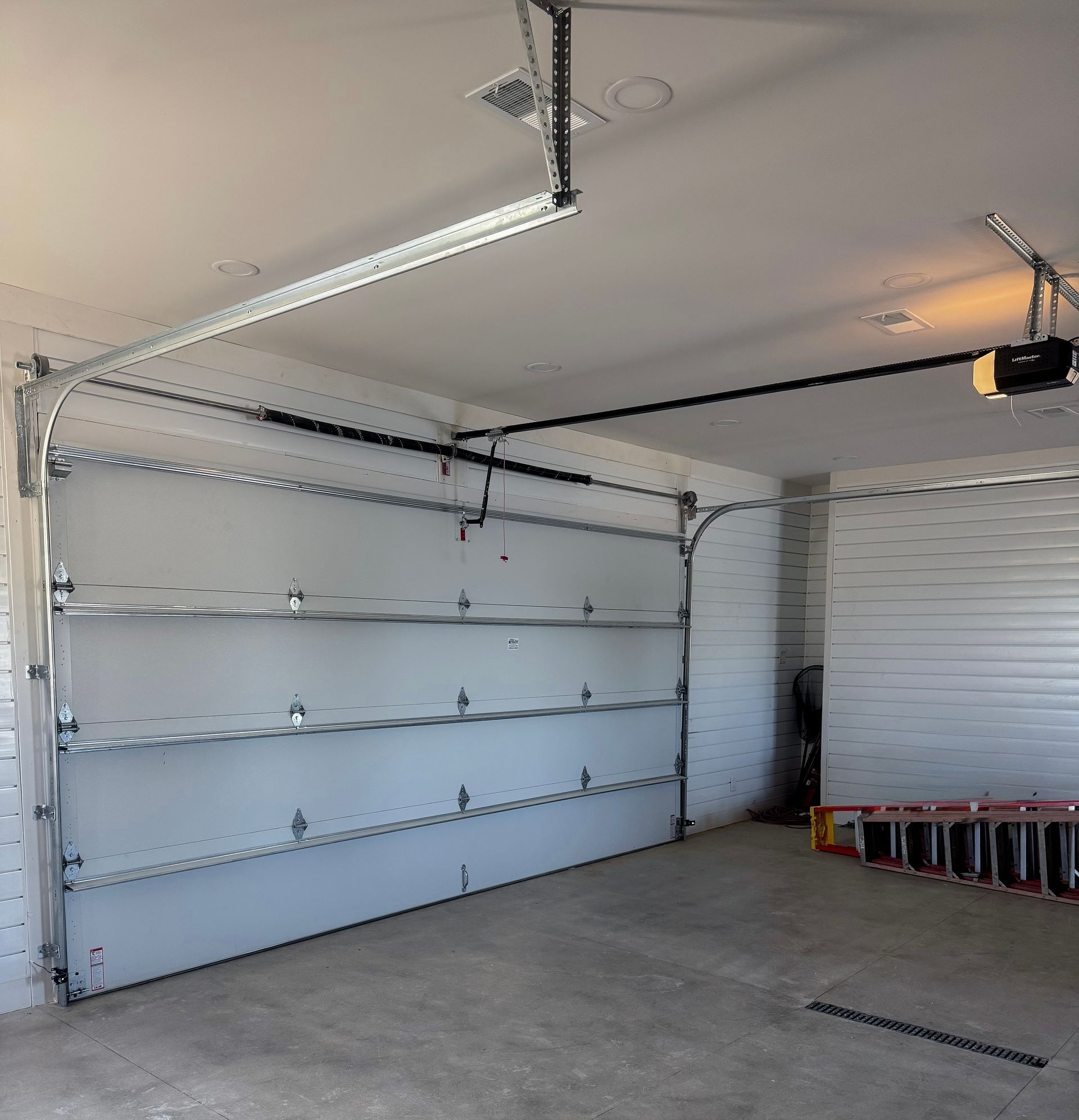 Empty garage interior with white paneled walls, ceiling track lights, and a ladder on the right.
