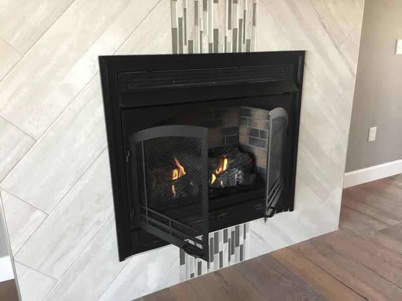 Modern black fireplace insert with glass doors on a light tiled wall, set above wood flooring.