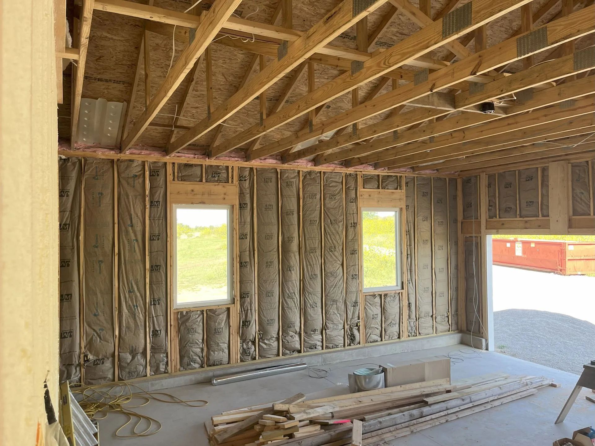 Framed interior of a house under construction with exposed studs, rafters, and bright window openings.