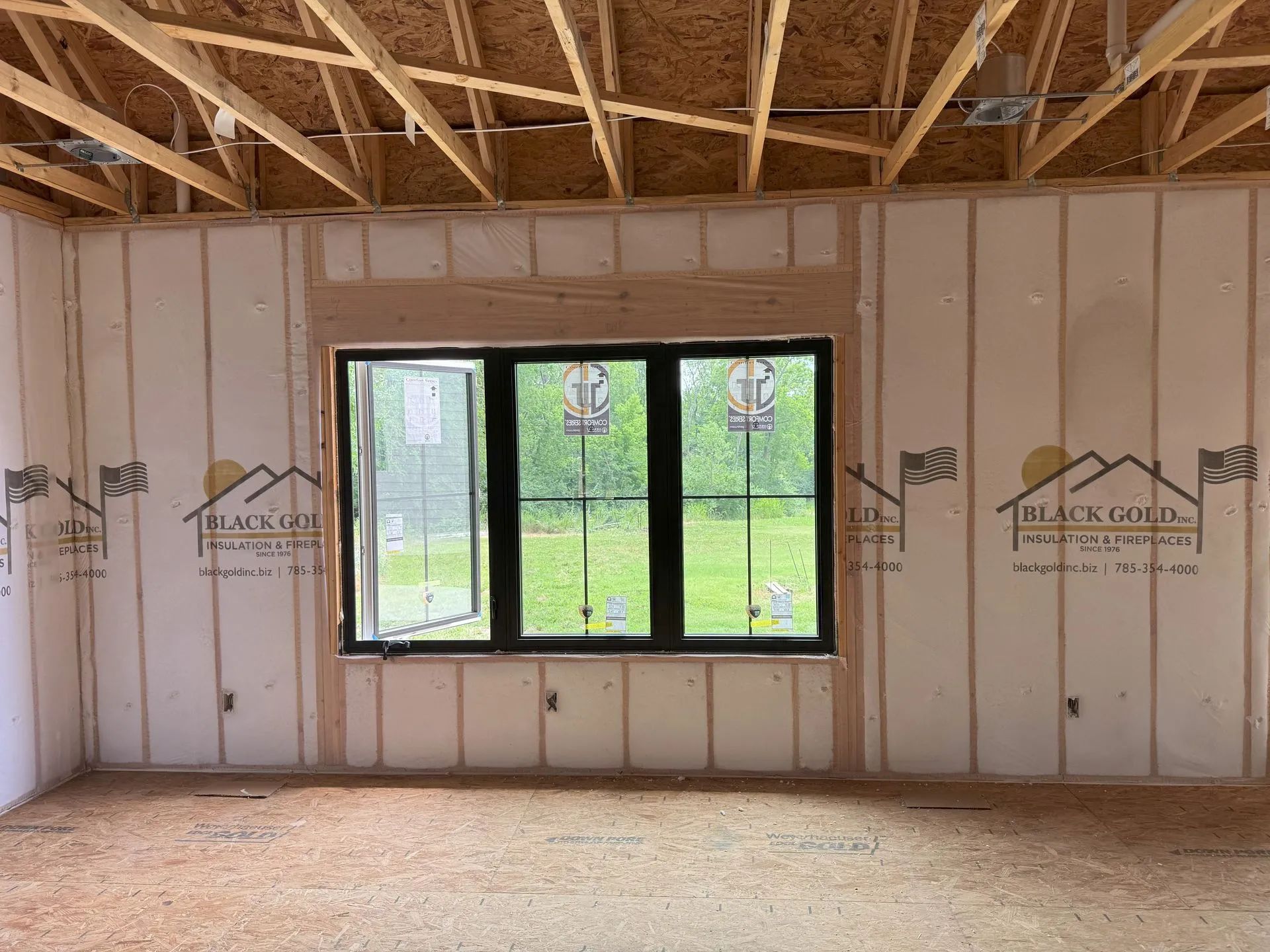 Unfinished room with exposed framing, white insulated walls, and a black-framed window overlooking green grass