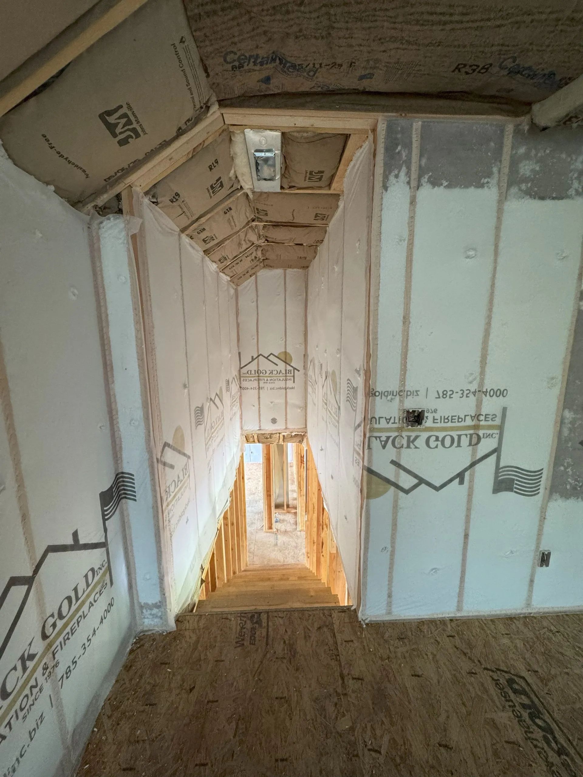 Unfinished stairwell with exposed framing, white insulation panels, and a wooden floor, viewed downward.