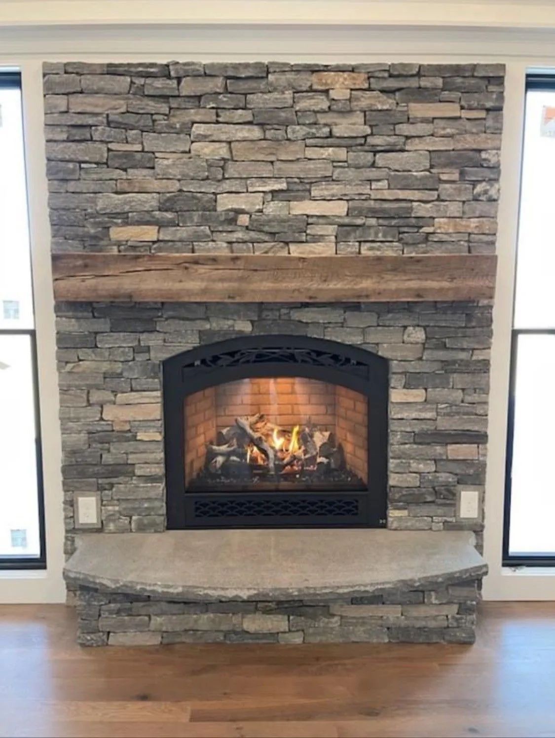Stone fireplace with lit fire and wooden mantel in a room with hardwood floors
