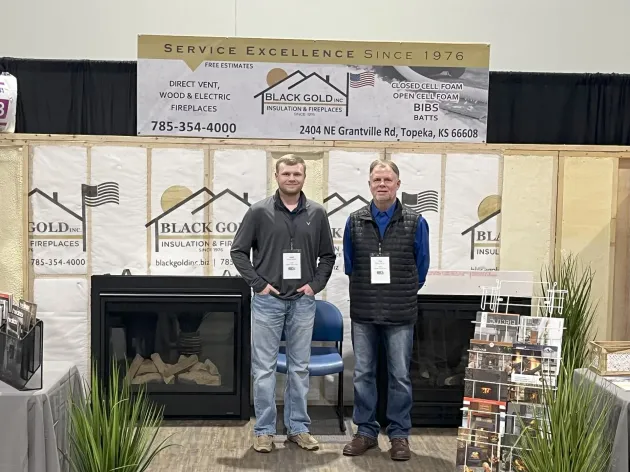 Two men standing in a booth display with a fireplace and product shelves at an expo.