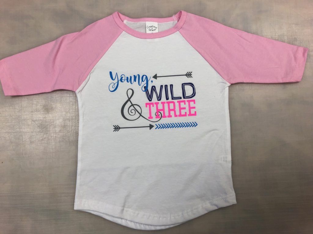 A pink and white t-shirt with the words `` young wild & three '' on it.