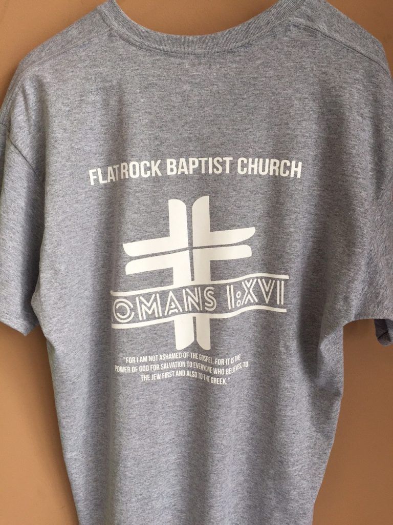 A gray t-shirt with a white cross on it is hanging on a wall.