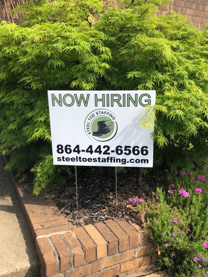 A now hiring sign is sitting on a brick wall next to a bush.