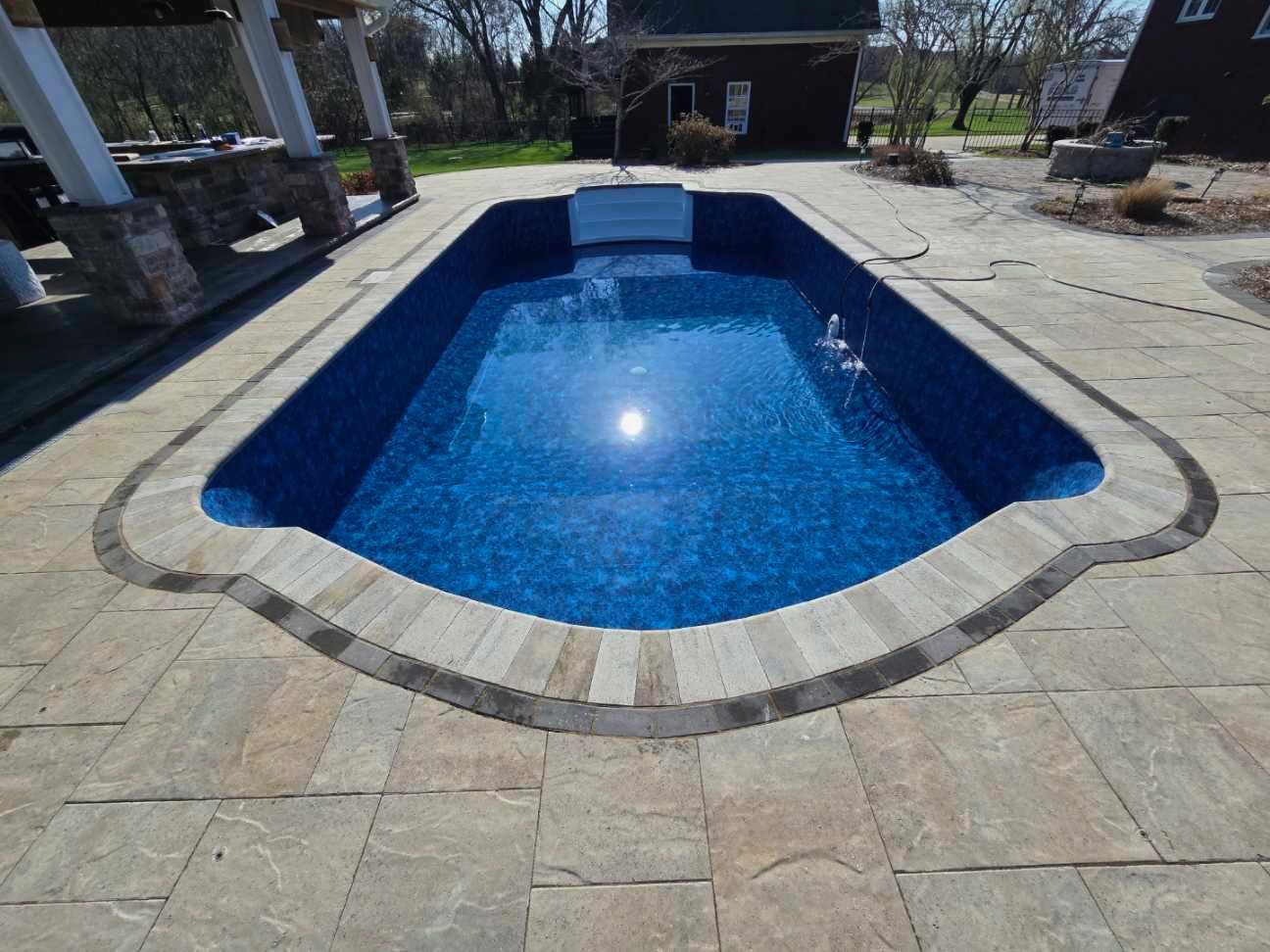 Rectangular in-ground swimming pool with blue mosaic tile and stone coping, surrounded by paved patio.