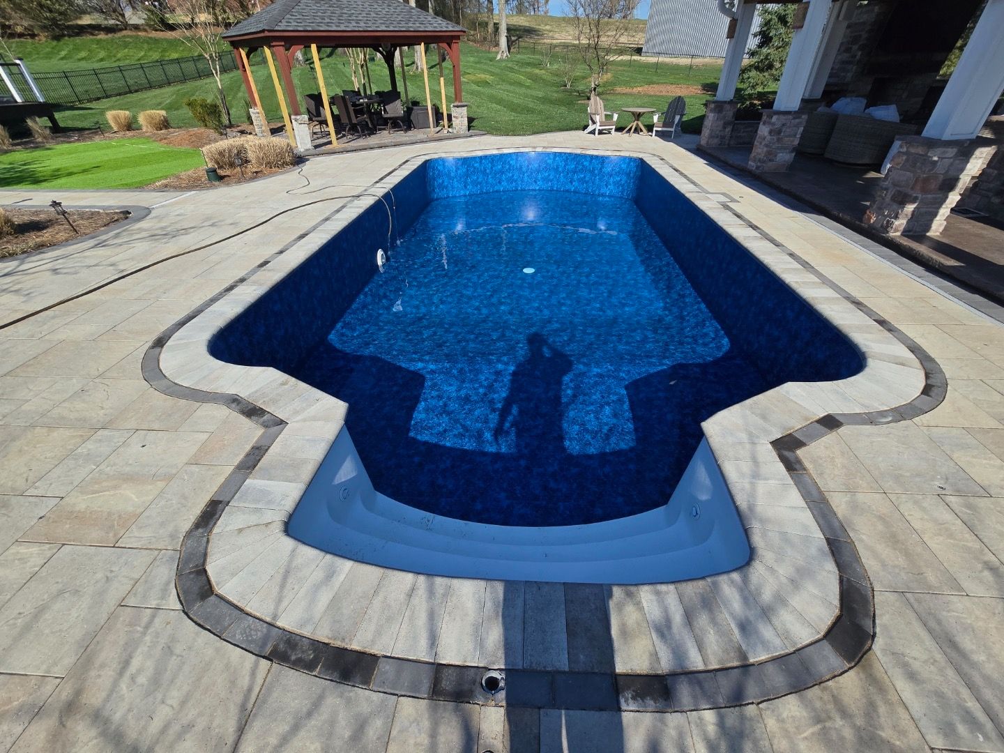 Swimming pool with blue water and steps, surrounded by stone patio. Gazebo and lawn in the background.