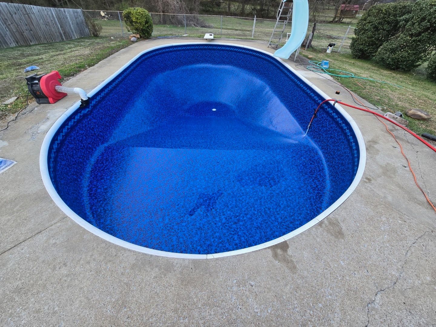 Oval-shaped in-ground pool, blue interior, surrounded by a concrete deck, red pump and slide visible.