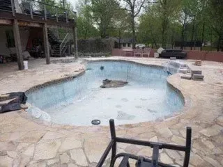 An empty kidney-shaped swimming pool undergoing renovation with surrounding stone patio and deck.