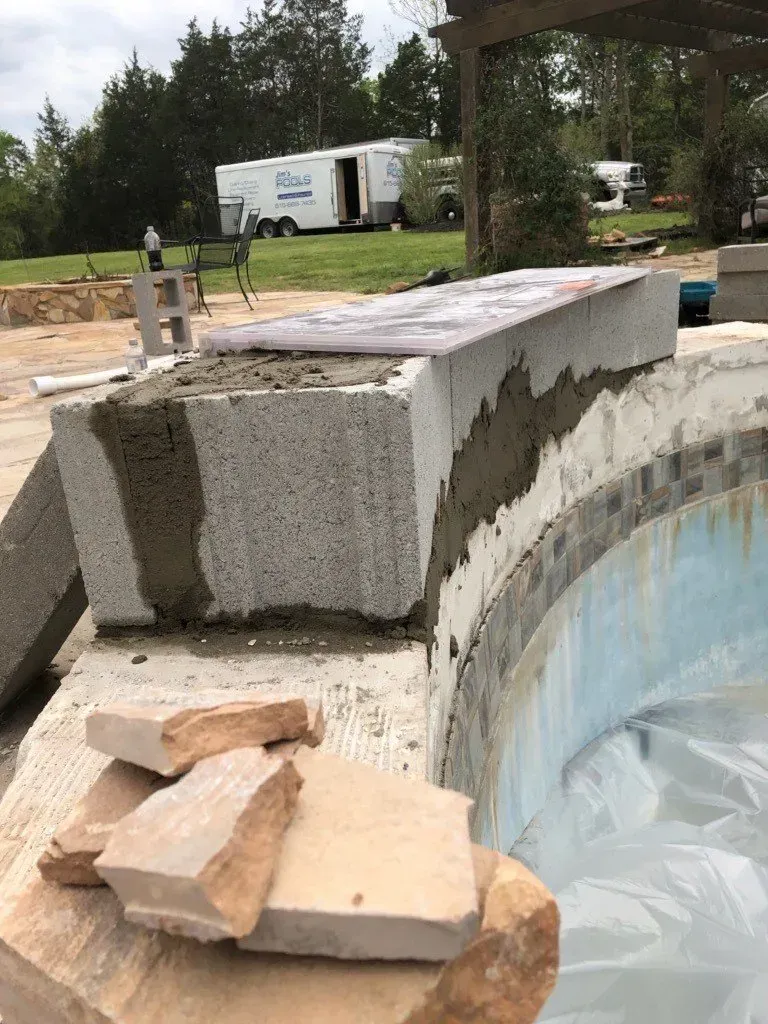 Construction of a pool coping; gray block and mortar set atop pool edge, tan stones nearby.