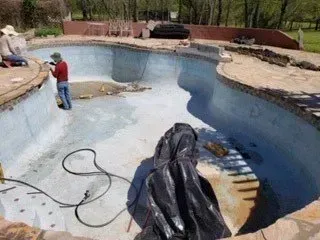 Workers renovating an empty, kidney-shaped swimming pool. The pool's surface is being repaired.