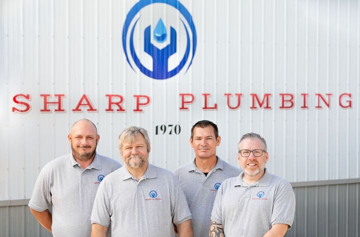 Four men in gray shirts stand in front of a building with the logo of Sharp Plumbing.