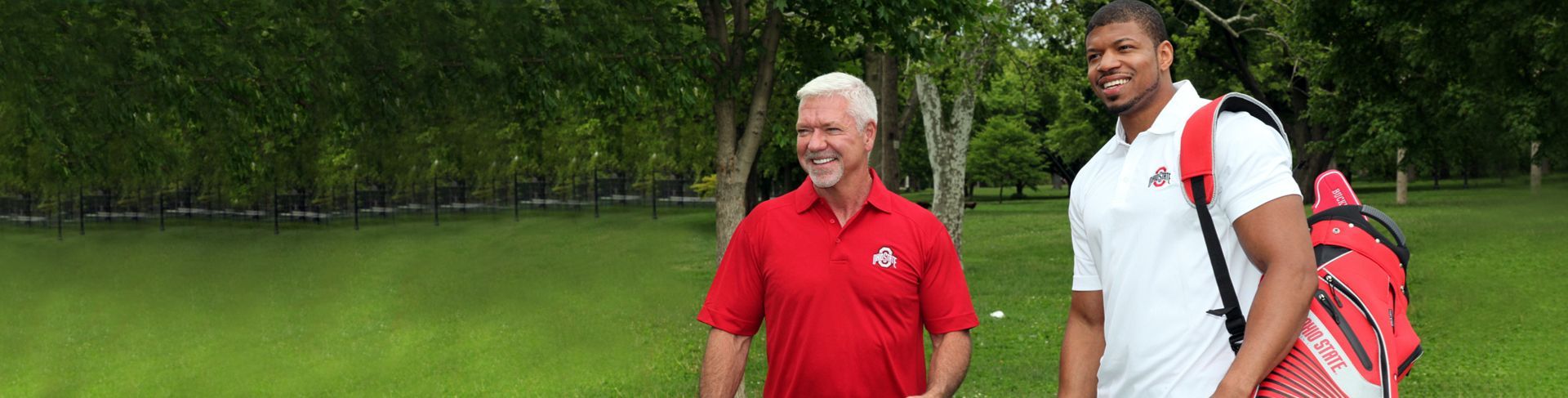 Two men are standing next to each other in a grassy field . one of the men is carrying a golf bag.