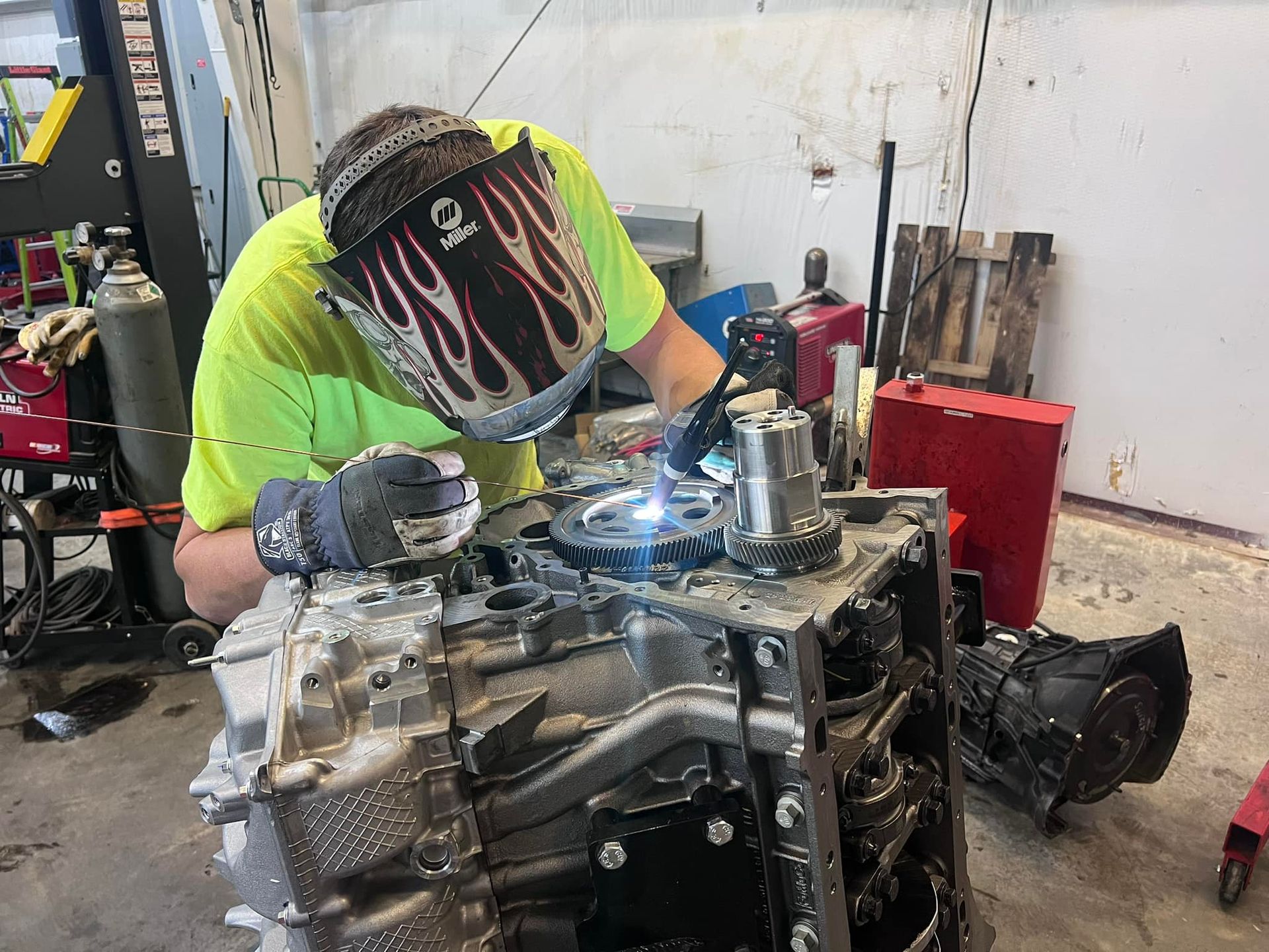 A man wearing a welding mask is working on a car engine.